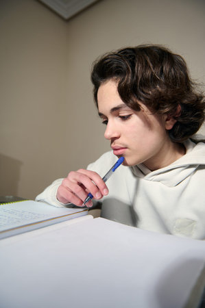 Teenage boy concentrating on homework at a cozy home desk in the evening holding a pen and notebook...