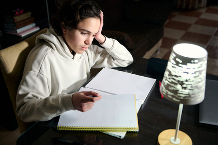Teenage boy studying at a desk in the evening under warm lamp light, in a cozy home interior looking...