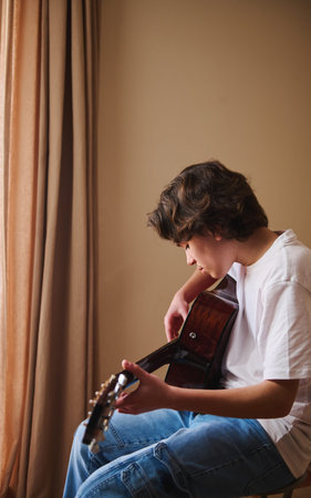 Teen boy sits by a window playing an acoustic guitar at home, focused on practice and learning music...