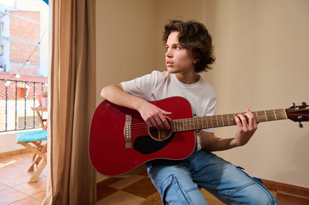 Teenage boy sits by a balcony window strumming a red acoustic guitar during a focused, casual...
