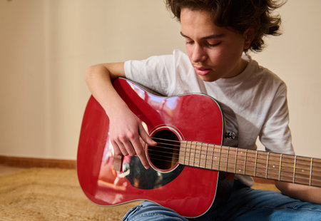 Teen boy sits on the floor at home concentrating as he plays a red acoustic guitar, practicing...