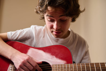 Teen boy concentrating while playing a red acoustic guitar indoors, practicing chords and learning...