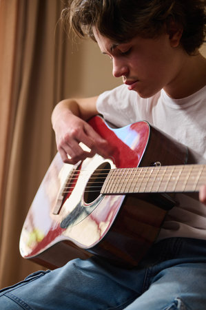 Teen boy focused while playing a red acoustic guitar at home, practicing chords and expressing calm concentration and creativity in a warm indoor setting.の写真素材