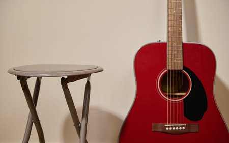 A red acoustic guitar leans beside a small folding stool against a beige wall, evoking a quiet practice or songwriting moment with warm tones and minimalist composition.の写真素材