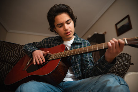 Teenage boy in casual clothes strums an acoustic guitar on a couch, practicing music in a warm, cozy...