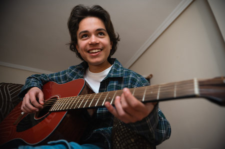 Smiling teenage boy in casual clothes plays an acoustic guitar in a cozy home interior, strumming...