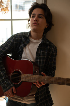 Teenage boy in a plaid shirt strums an acoustic guitar by a window, practicing music at home with a...