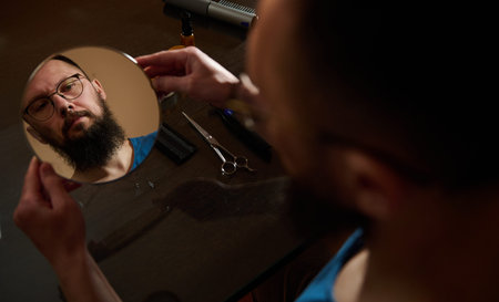 Bearded man with glasses studies his reflection in a small mirror on a table with scissors, comb and grooming oil, a quiet moment of personal care and preparation to trim facial hair.の写真素材