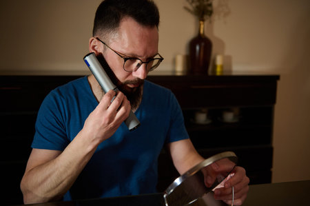 Bearded man wearing glasses grooms his facial hair with an electric trimmer while checking progress...
