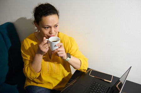 Female entrepreneur and digital nomad sips freshly brewed coffee in a cozy cafe while telecommuting...