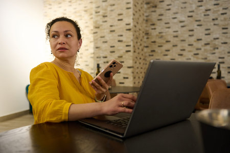 Female entrepreneur and digital nomad in a cozy cafe using a laptop and smartphone while enjoying...