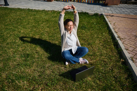 Young woman seated on grass stretching arms while using a laptop outdoors, enjoying fresh air,...