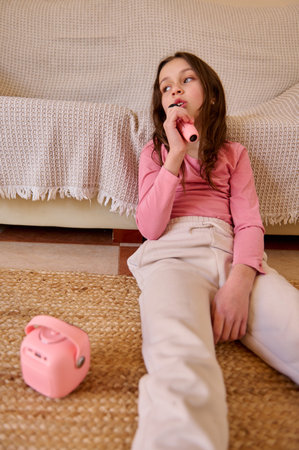 A relaxed young girl sings into a pink microphone while sitting on the living room floor beside a...