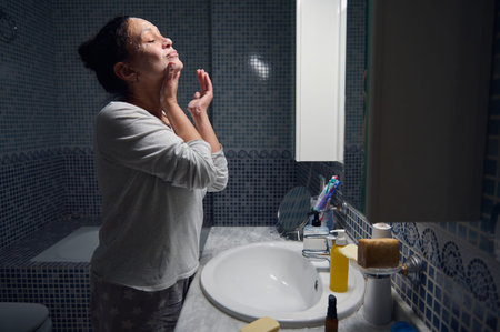 Middle-aged woman in pajamas gently washing her face at the bathroom sink during a morning skincare...