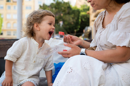 A toddler opens his mouth to eat a watermelon piece offered by a caregiver during a summer snack break on a wooden bench.