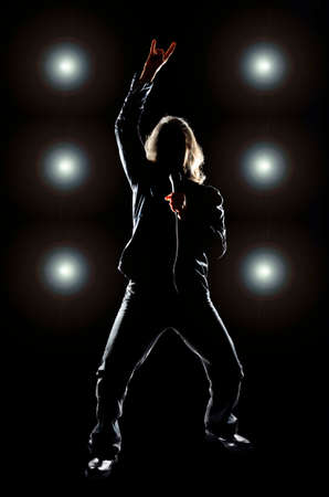 Man with long hair holding a microphone, against a black background with lights behind him Man with long hair holding a microphone, against a black background with lights behind him