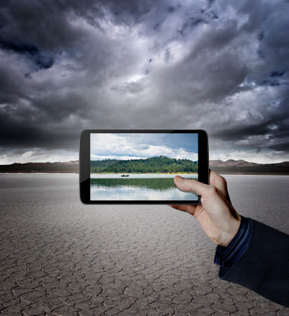 Hand holding a digital tablet in a desert with a lake on the screenの写真素材