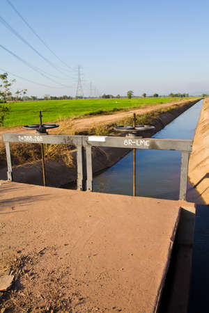 Water gate manal wheel control of irrigation canal with farmland and high voltage pylonの写真素材