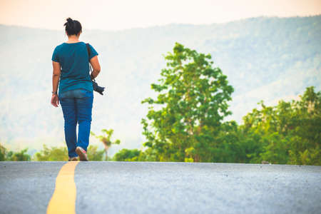 Woman with camera on shoulder walking on the road in countrysideの写真素材