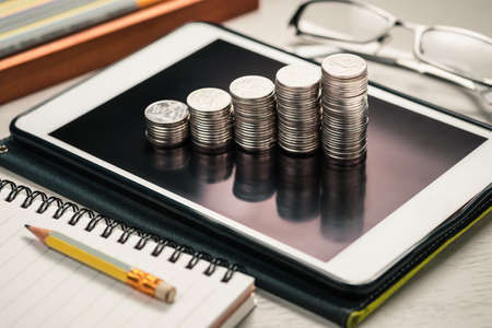 Turn Idea to income, closeup heap coins stair arranged on mobile tablet with writing tools on the deskの写真素材