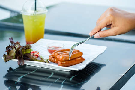 Closeup hand stab the sausages with fork on the white plate, afternoon appetizer sausages served with salad and iced juice on the tableの写真素材