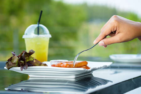Closeup hand stab the sausages with fork on the white plate, afternoon appetizer sausages served with salad and cold drink on the tableの写真素材