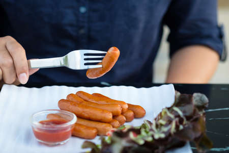 Closeup hand stab the sausages with fork over the white plate, afternoon appetizer sausages served with ketchup and saladの写真素材