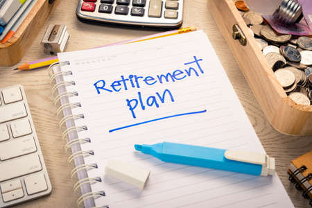 Closeup Retirement Plan text by handwriting on notebook with part of coins in the wood box,...