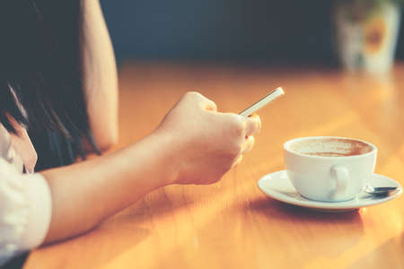 Closeup woman hand using smartphone with cup of coffee on the table, phone and coffee addict as modern usual lifestyle conceptの写真素材