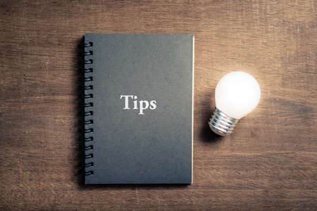 Black notebook with text TIPS and glowing light bulb on wood backgroundの写真素材