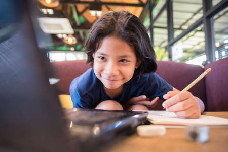 Child girl sit on the sofa, watching the screen on laptop and going to write a lesson on notebook, online education at homeの写真素材