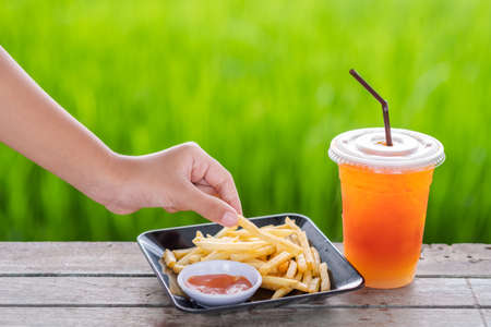 Closeup hand take the french fries from the dish that served on the outdoor table near the green field with lemon iced teaの写真素材