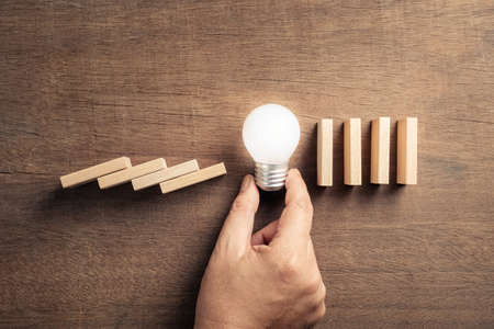 Hand place a glowing and powerful light bulb in the row of wood block set as domino falling symbol, to stop and prevent the collapse, creative and powerful solutionの写真素材