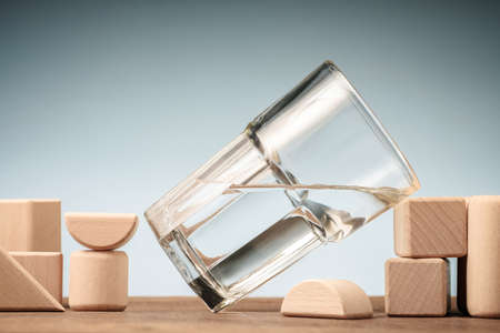 Titled glass of water with half full water lean on the wood block toy on the table, glass half full conceptの写真素材
