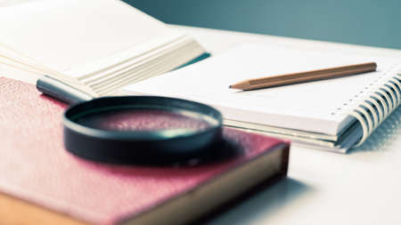Closeup pencil on notebook with part of opened book and magnifying glass, reading for writing,...