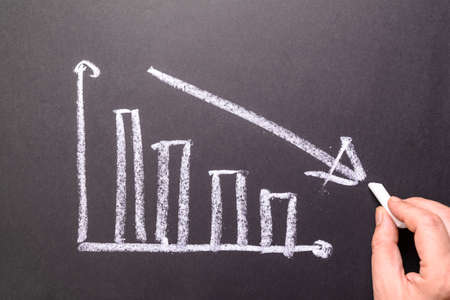 Hand sketching decreasing graph on chalkboard, business going down, decrease sales and revenue in economic crisisの写真素材