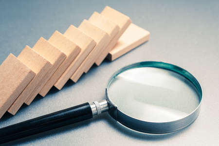 Magnifying glass and a row of fallen domino on gray background, domino effect analysis conceptの写真素材