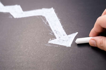 Closeup hand draw an arrow line that tends to go down on the chalkboard, metaphor to business or investment, recessionの写真素材