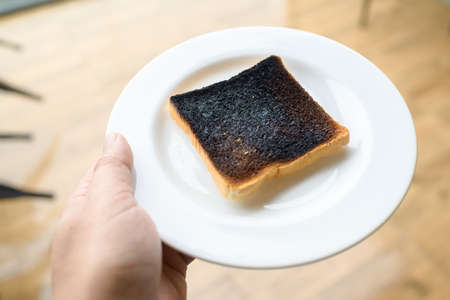 Hand holding the white dish with burnt toast on it, over heat, and mistake in the morning, danger of charred foodの写真素材