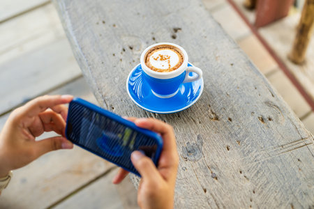 Closeup a woman is going to take a picture of cappuccino coffee on the wood bench with smart phoneの写真素材