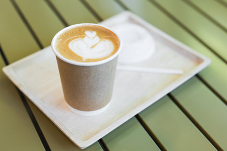 Cappuccino coffee in takeaway paper cup, served on the wood tray and green tableの写真素材