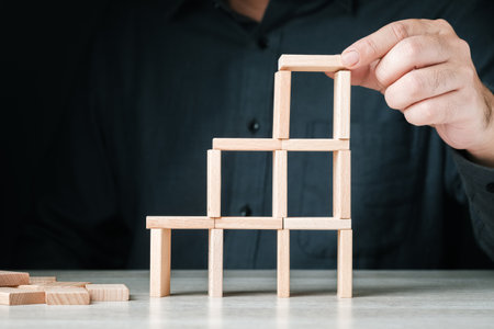 A man build a tower with small wooden pieces, try to increase the level, but looks risky to fall down, fragile, easy success, or insurance conceptの写真素材