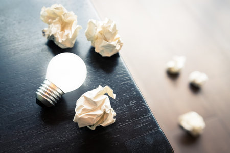 Closeup glowing light bulb with some crushed paper balls on the desk, and fallen on the ground,...