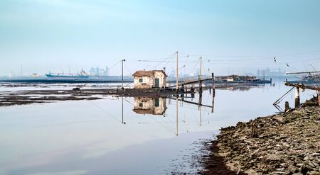 Old fishing house reflecting in the lagoon of Marina di Ravenna, industrial harbour on background.