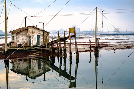 Old fishing house reflecting on the lagoon of Marina di Ravenna, industrial harbour on background.の写真素材