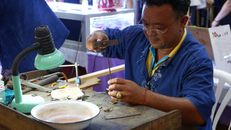 BANGKOK, THAILAND - 10 JULY, 2019: Ethnic jeweler working during exhibition. Ethnic craftsman in glasses sitting at shabby table and making jewelry while taking part in trade show.のeditorial素材