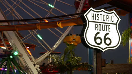 SANTA MONICA, LOS ANGELES, USA - OCT 28, 2019: Iconic road sign glowing, historic route 66. Famous california symbol, pier of pacific ocean resort. Illuminated festive ferris wheel in amusement park.のeditorial素材