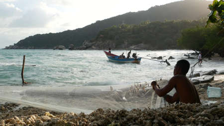 KOH PHANGAN, THAILAND - 11 JANUARY 2019 Ethnic fisherman hanging bait on net. Side view of ethnic shirtless man hanging small fish on net while sitting on shore near sea.のeditorial素材