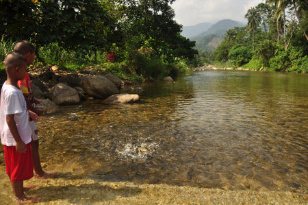 NAKHONSITHAMMARAT, THAILAND - JUNE 7, 2014: Kids in shallow clear water of pond, View of little ethnic boys standing in clear shallow water of flow with fish splashing. Mountain on backgroundのeditorial素材
