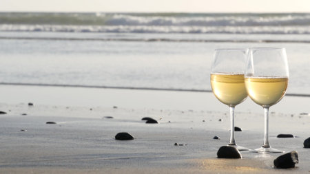 Two wineglasses on sandy ocean beach. Couple of glasses and pebbles, white wine for romantic date near sea water. Cozy beachfront sunset near tide waves. Seamless looped cinemagraph. California, USA.の写真素材